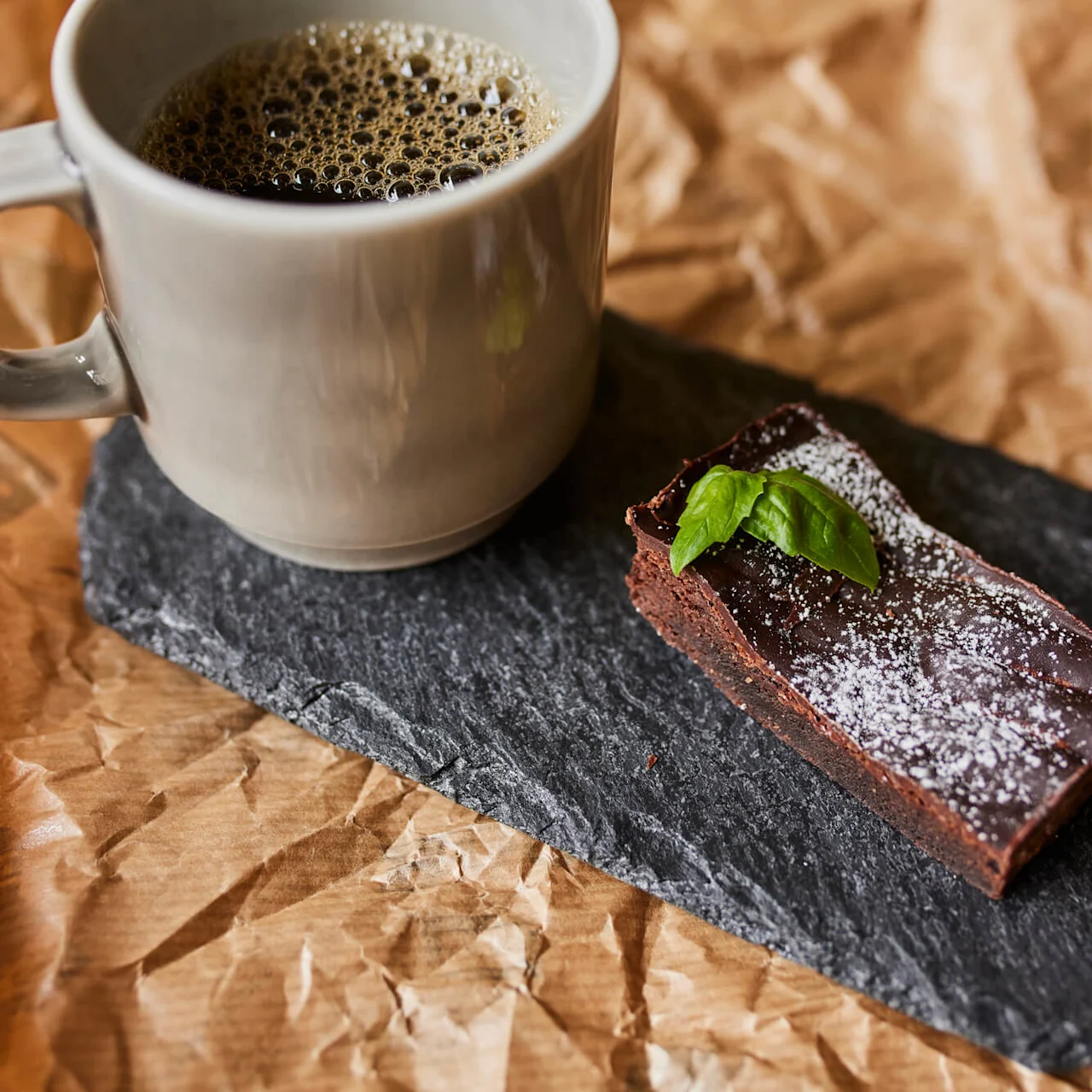 En kopp svart kaffe står ved siden av en sjokoladebrownie pyntet med melis og et basilikumblad, plassert på en skiferplate over krøllet brunt papir.