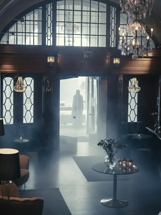 En svagt oplyst, elegant lobby med en lysekrone, sofaer og et centralt bord med blomster og lys. En silhouet står ved den lyse indgang.