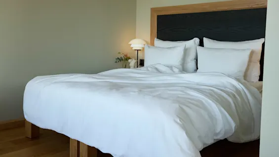A neatly made bed with white linens sits against a wooden headboard. A bedside lamp glows softly on a nightstand with a small vase of flowers.
