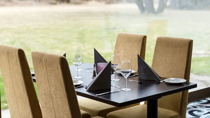 A dining table is set with wine glasses, folded napkins, and plates surrounded by beige upholstered chairs. It overlooks a grassy field through large windows.