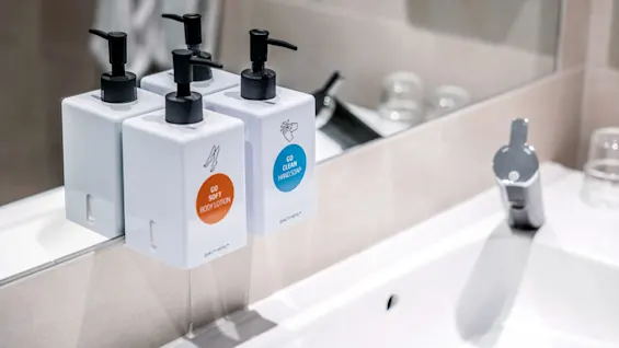 Tre vita dispenserflaskor med svarta pumpar märkta "GO SOFT BODY LOTION" och "GO CLEAN HAND SOAP" är monterade mot en spegel ovanför ett vitt handfat.