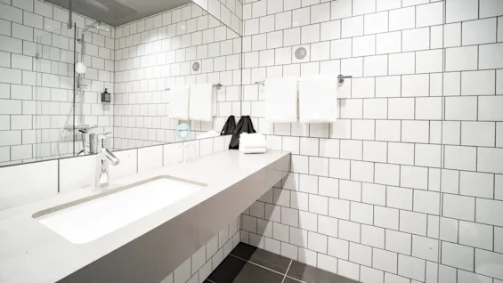 Moderni kylpyhuone, jossa tyylikäs pesuallas, kaksi taitettua pyyhettä hyllyllä, suihkualue ja laatoitettu seinä. Kyltti "DRINK WATER" lasin vieressä.