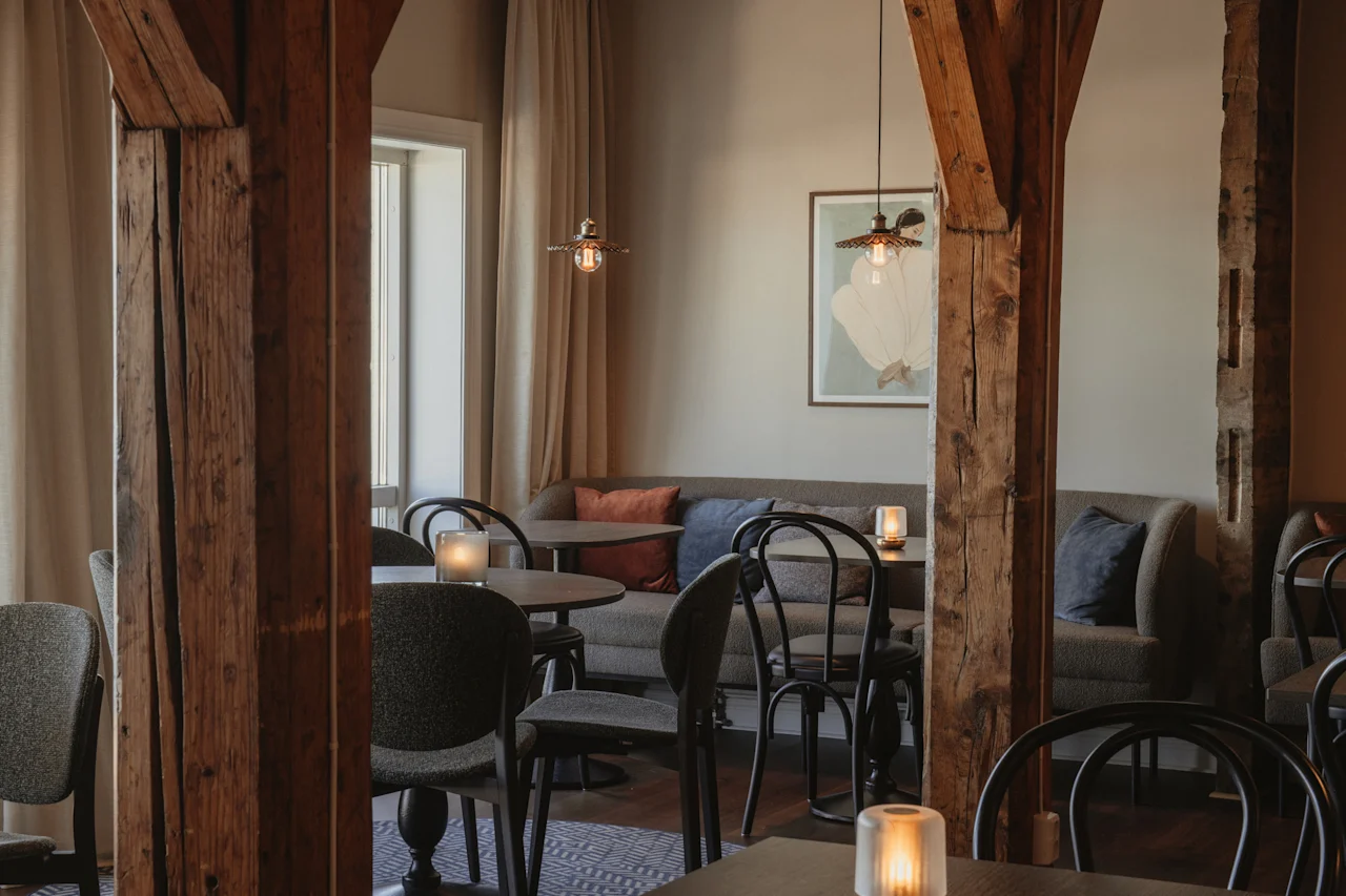 Et varmt og indbydende spise- eller loungeområde med træbjælker, komfortable siddepladser og blød belysning fra hængende lamper og stearinlys.