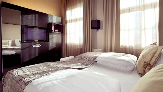 Et moderne hotellrom med to senger, skarpt hvitt sengetøy og sollys som filtreres gjennom tynne gardiner.