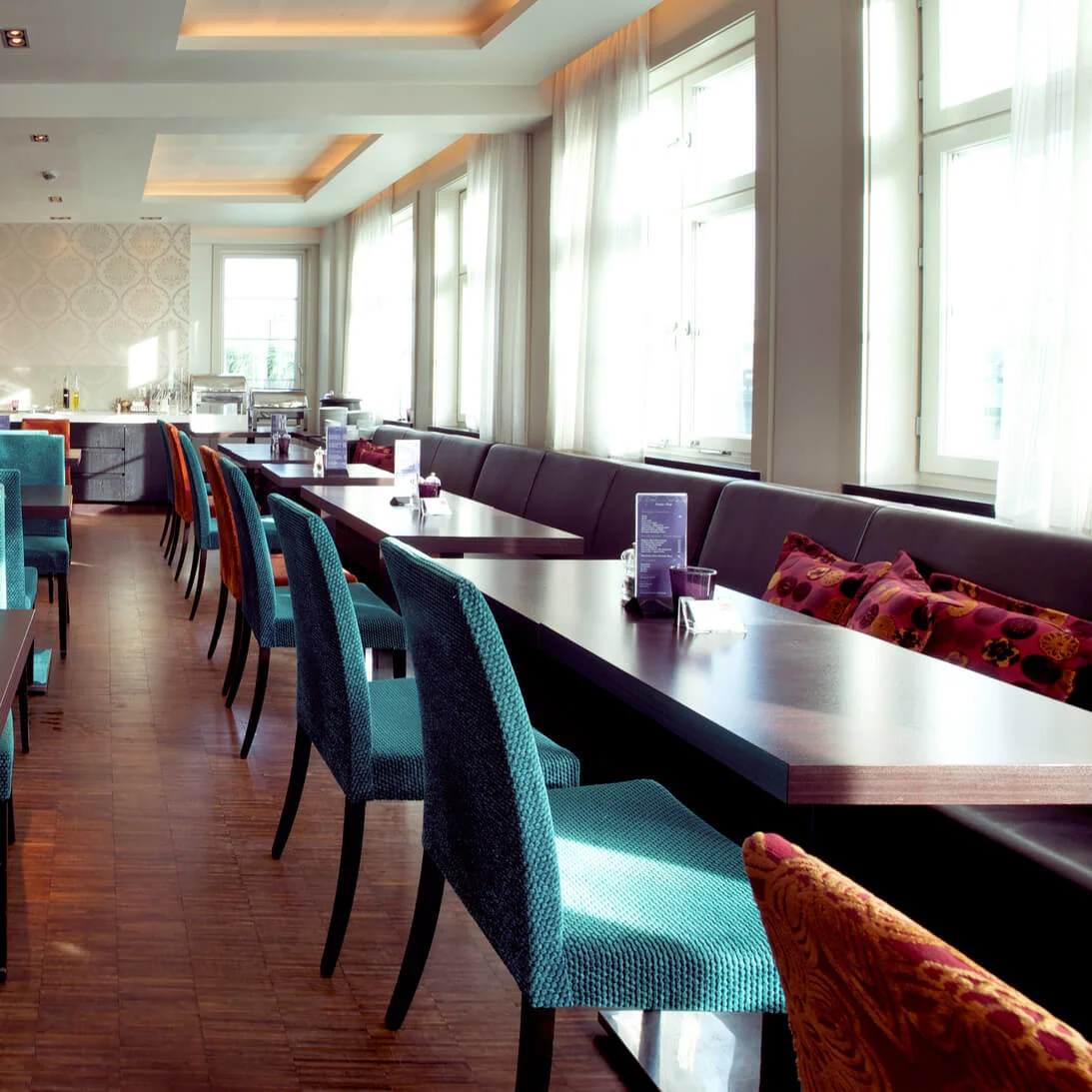 Et tomt, moderne restaurantinteriør med fargerike polstrede stoler, trebord og store vinduer som slipper inn dagslys.