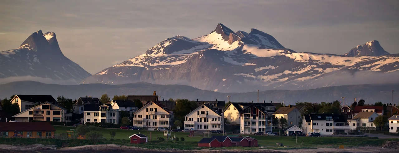 Snötäckta berg står majestätiskt under en molnig himmel och blickar ner över en liten kuststad med hus och röda stugor vid vattnet.