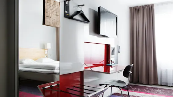 En modern hotellrum har en säng med beige huvudgavel, en väggmonterad TV, rött skrivbord med stol och ett fönster täckt med gardiner. En brun papperspåse hänger från en klädställning.