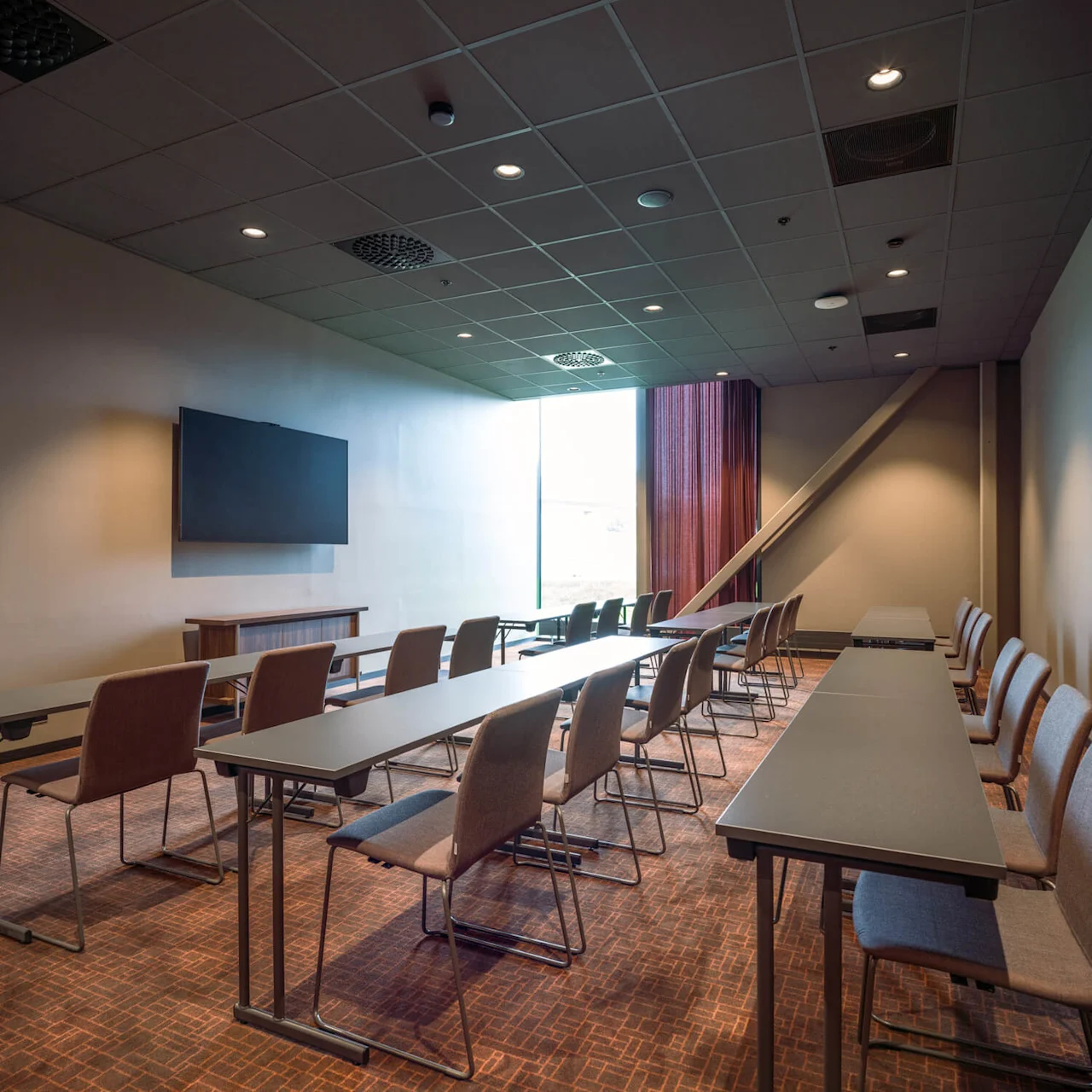 Tomt konferanserom med rader av stoler og bord vendt mot en stor veggmontert skjerm. Omgivende belysning fra taklamper; et stort vindu med gardiner lyser opp rommet.