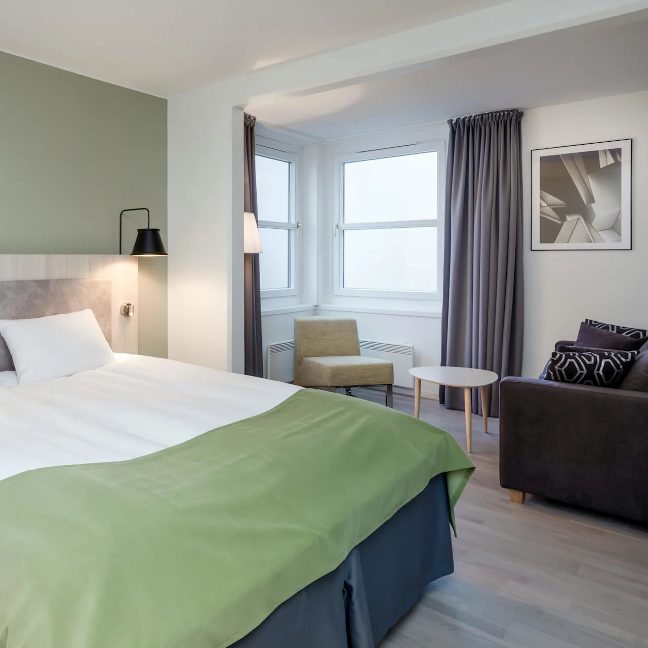 Bed with white sheets and green throw sits against light green wall, flanked by black lamps. A chair and table populate the room's corner by curtained windows.