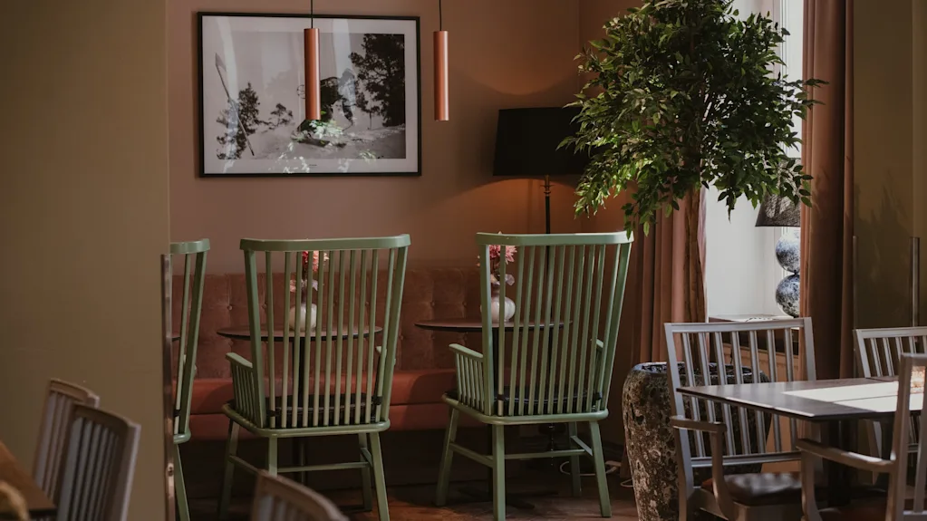 Et varmt og innbydende spiseområde med lysegrønne stoler, en stor plante og et innrammet kunstverk på veggen, badet i naturlig lys.