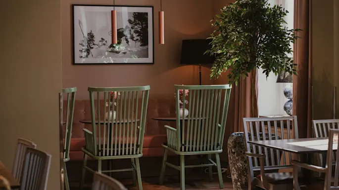 Et varmt og innbydende spiseområde med lysegrønne stoler, en stor plante og et innrammet kunstverk på veggen, badet i naturlig lys.