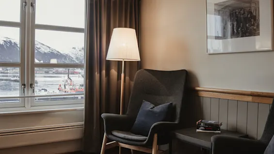 A cozy seating area with an armchair, lamp, and a window view of snowy mountains and water.