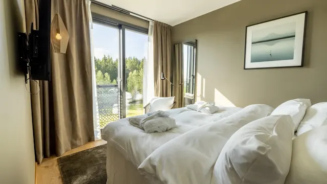En pent oppredd seng med hvitt sengetøy er i et rom med brune gardiner som avslører glassdører ut mot en balkong med utsikt over trær. Kunstverk henger på den grønne veggen.