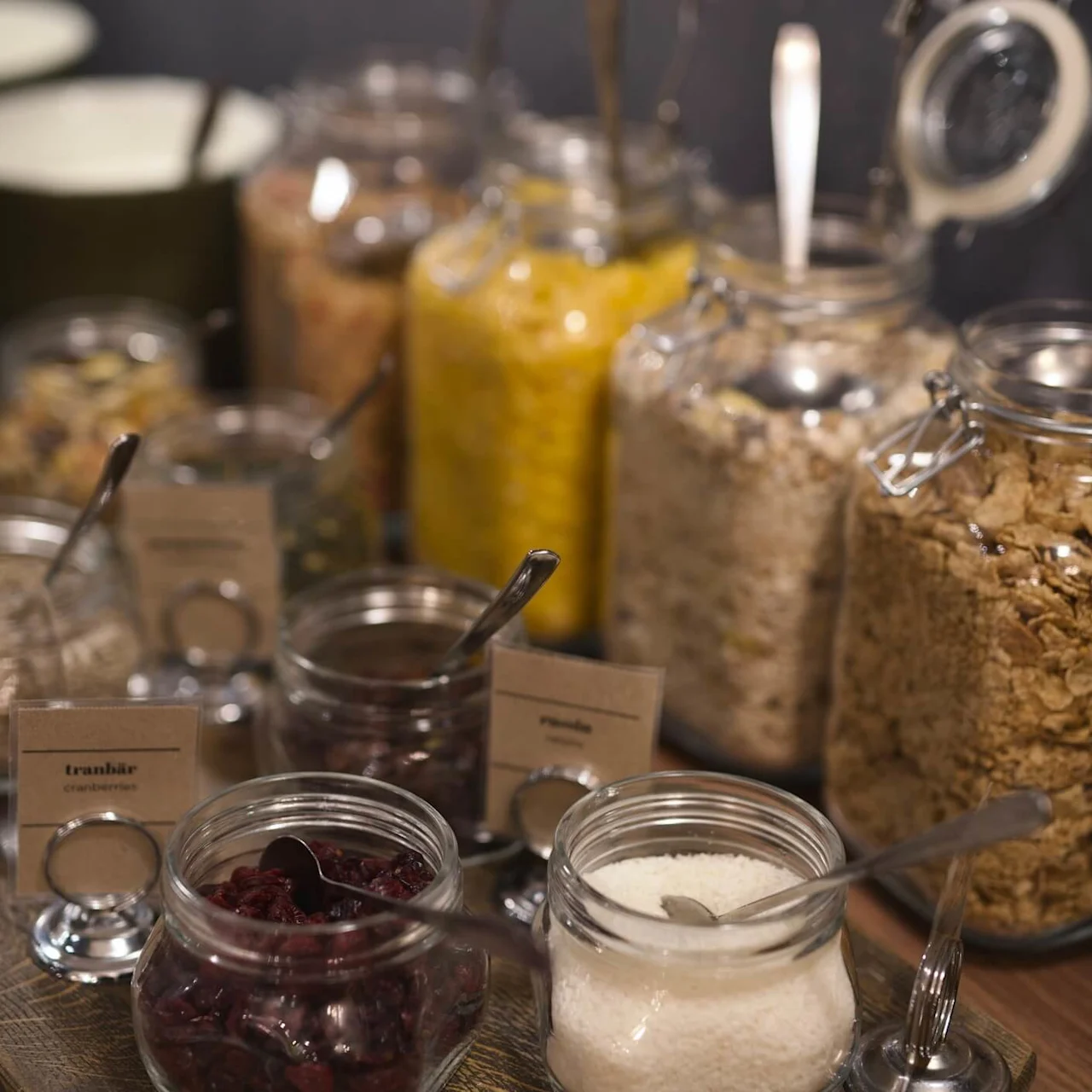 Glass jars with ingredients for breakfast, labeled with tags, against a dark backdrop. Visible tags read 'tranbär' (cranberries) and 'frukostflingor' (breakfast cereals).
