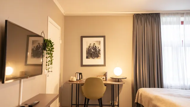 TV montert på veggen viser en refleksjon; skrivebord med vannkoker og lampe under innrammet fotografi; tilstøtende seng og gardiner i et mykt opplyst rom.