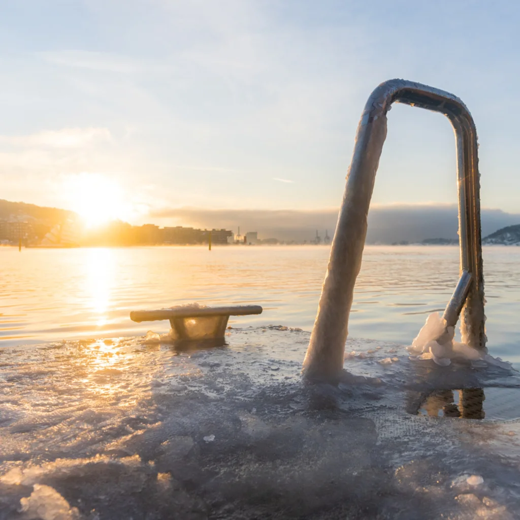 En fredfyldt vintermorgen ved vandet, med en iset stige der inviterer til en forfriskende dukkert, mens solen står op over byen.