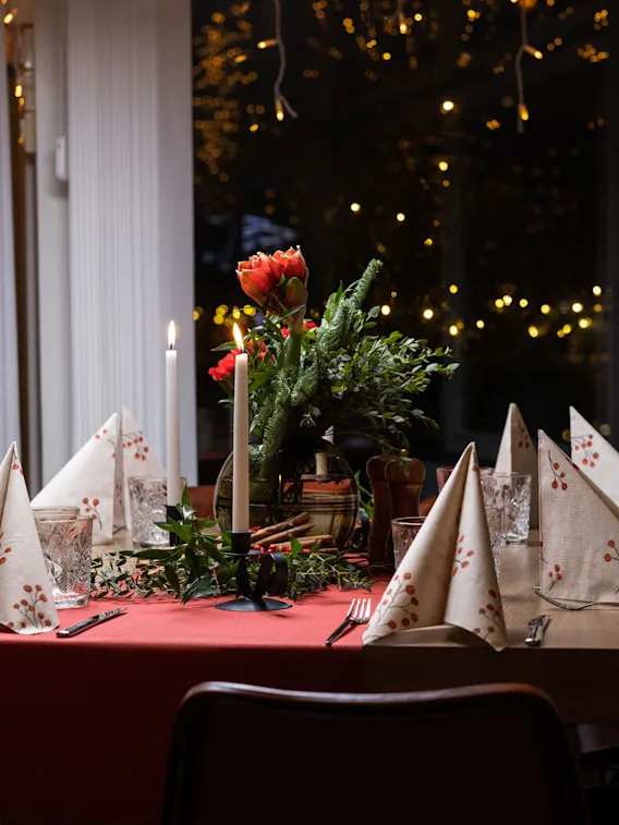 A warmly lit dining table with a floral centerpiece, candles, and neatly folded napkins, against a backdrop of twinkling lights.
