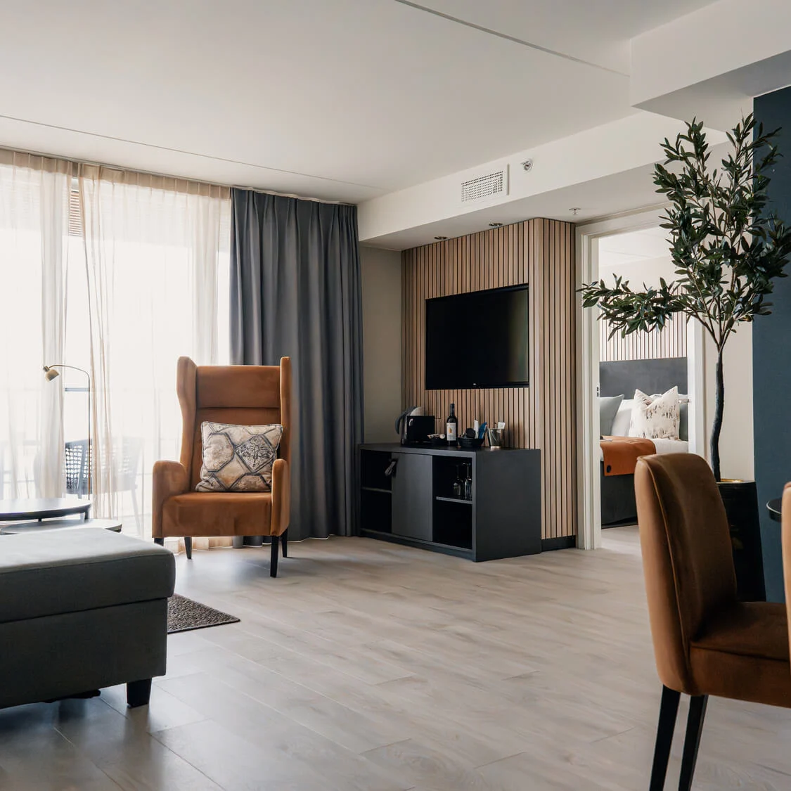 Orange lenestol står ved et kaffebord i et lyst rom med gulv-til-tak-vindu og gardin. En sofa, TV og inneplante er til stede.