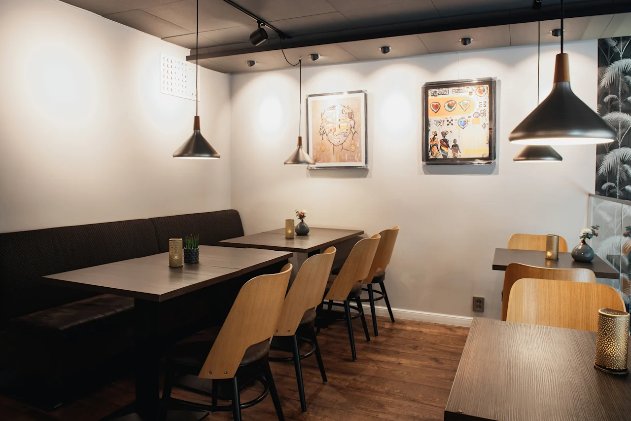 Et hyggeligt restaurantinteriør med mørke træborde, lyse træstole og en lang bænk. Pendellamper oplyser rummet, og kunstværker pryder væggene.