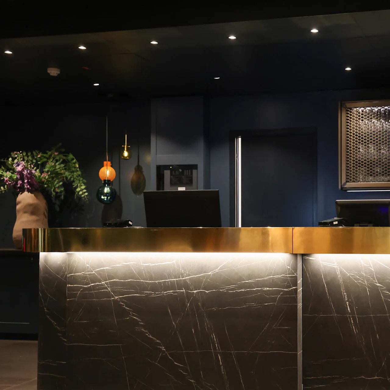 A reception desk with two computer monitors, set against a blue backdrop under dim lighting, with drinks displayed in a mesh cabinet and decorative plants nearby.