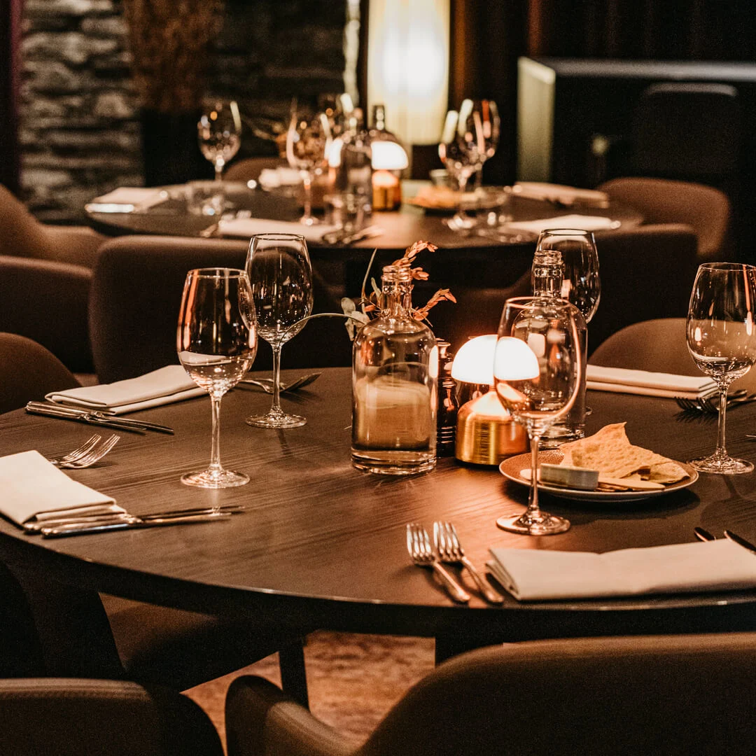 Elegantly set round dining table with wine glasses, cutlery, and candles, evoking a warm, intimate atmosphere in a refined restaurant.