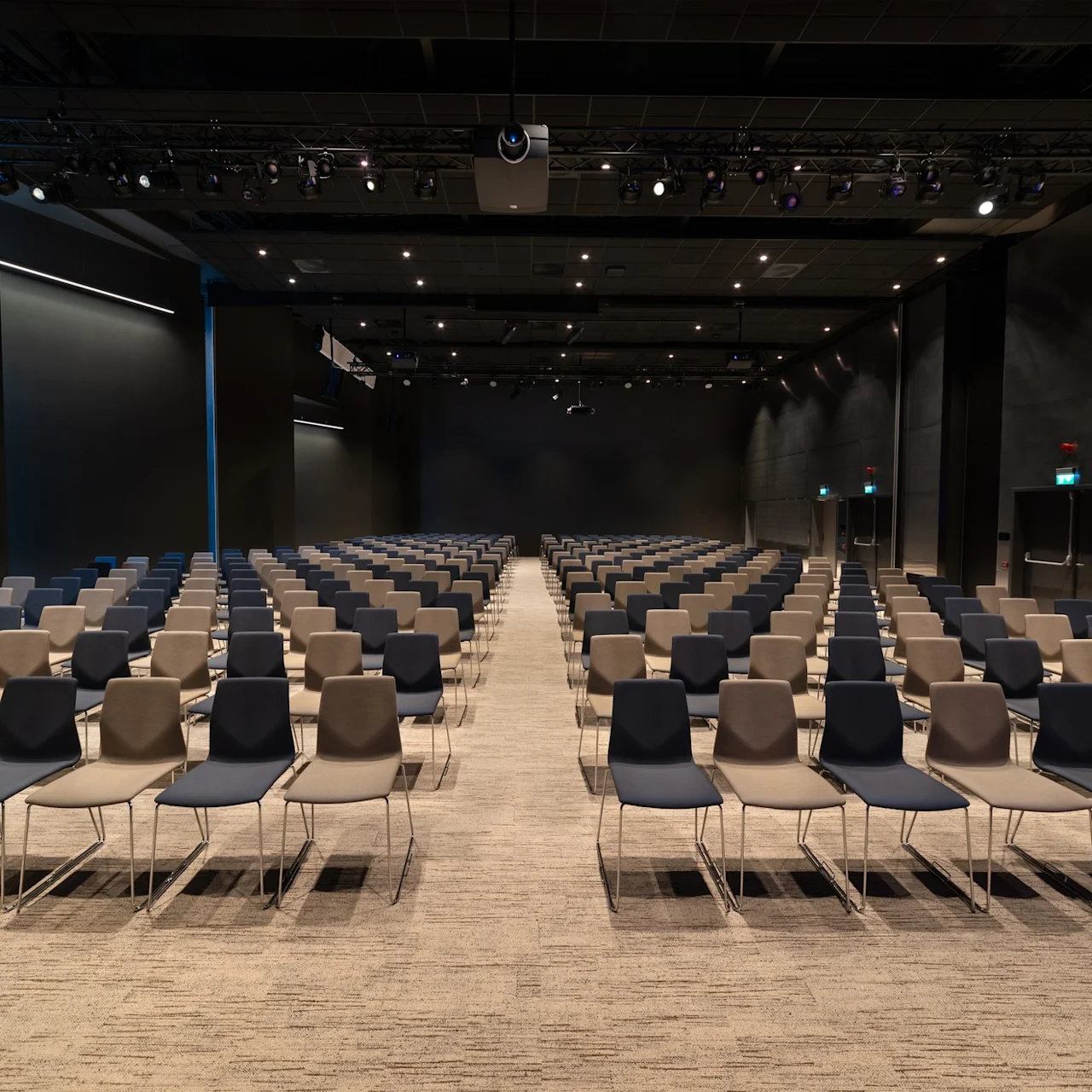 Rader med tomme stoler vendt fremover i en stor, svakt opplyst konferansesal, med taklys og utgangsdører på sidene.