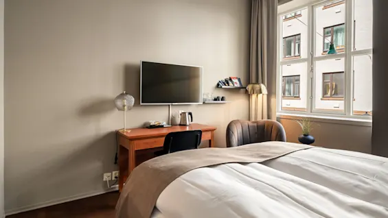 Et skrivebord med et TV på væggen står i et pænt hotelværelse. I nærheden er en stol, seng og et vindue med udsigt over tilstødende bygninger.