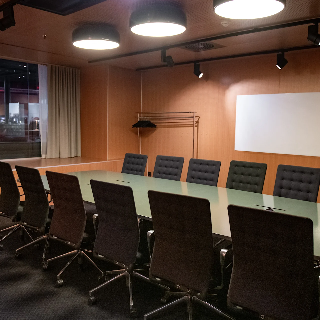 Ett långt glaskonferensbord omges av svarta kontorsstolar i ett träpanelerat rum med ett stort fönster, en whiteboard och taklampor.