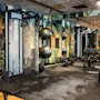 Fitnessudstyr, placeret i et moderne fitnesscenter, inkluderer håndvægte på stativer, træningsmaskiner og træningsbolde mod en farverig geometrisk-mønstret væg, der skaber en energisk træningsstemning.