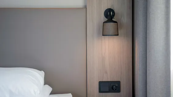Seng med hvide puder ved siden af et natbord. Væghængt sort lampe lyser mod et lyst træpanel ved siden af et gardin, hvilket skaber en minimalistisk og rolig soveværelsesindretning.