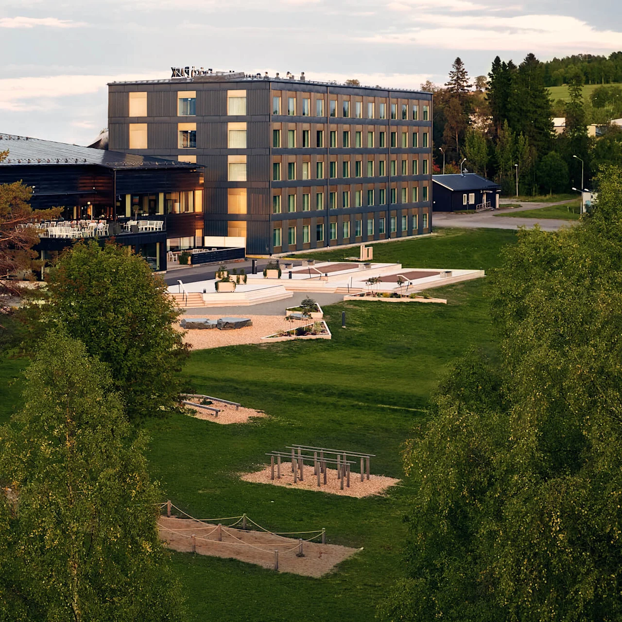 En modernt fler-våningshus med stora fönster ligger intill en utomhusterrass. Omgiven av frodiga gröna träd och öppna gräsytor under en delvis molnig himmel.