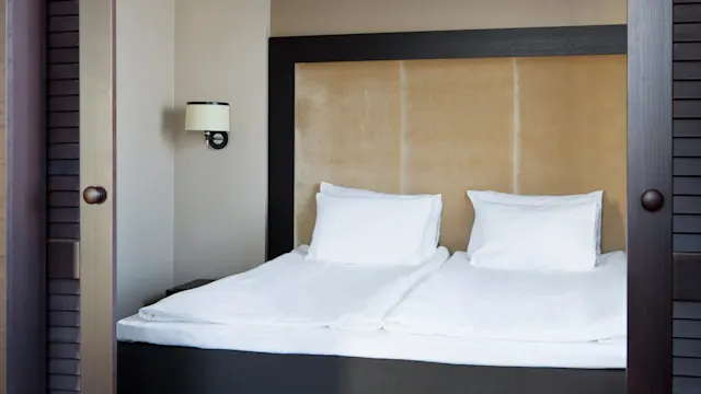 Bed with white sheets and pillows, framed by a dark wooden headboard, set in a beige room with a wall-mounted light fixture on the left.