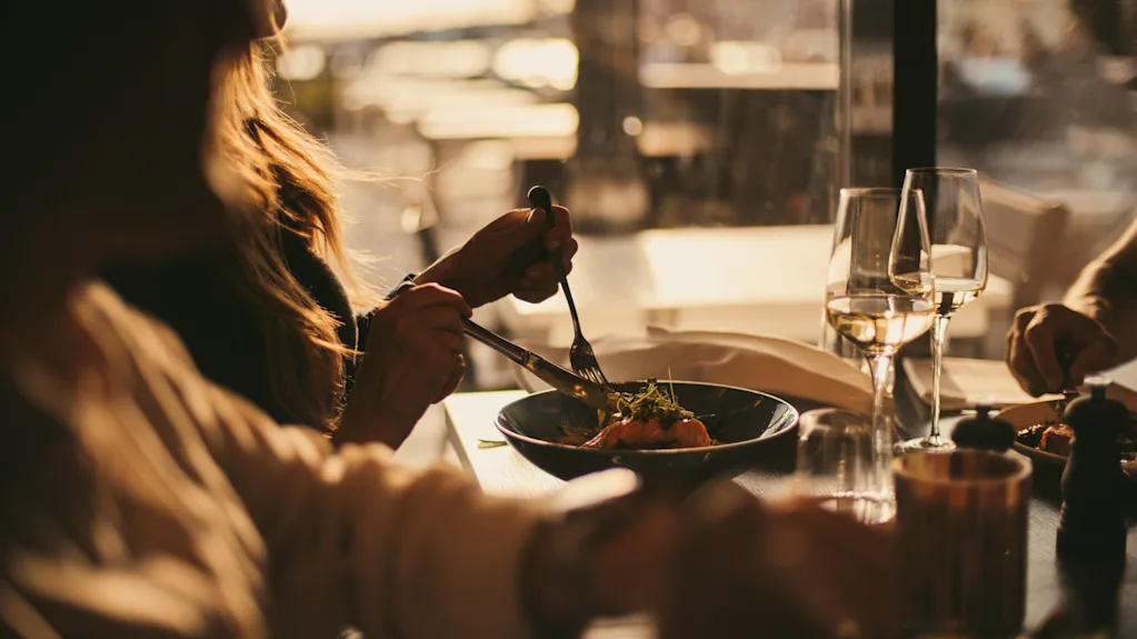 En person nyder et måltid med vin på en restaurant, med varm belysning og en sløret udendørsudsigt i baggrunden.