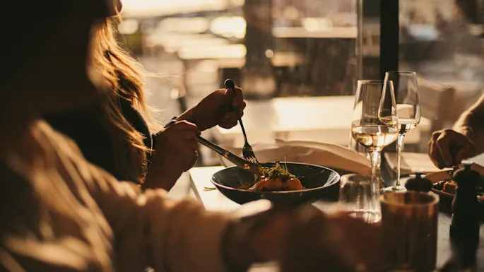 En person avnjuter en måltid med vin på en restaurang, med varm belysning och en suddig utomhusvy i bakgrunden.