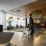 En person spaserer gjennom en elegant hotellobby med en hund, omgitt av frodige planter og moderne sitteplasser. Varme pendellamper skaper en koselig atmosfære, med geometriske mønstre på gulvet.