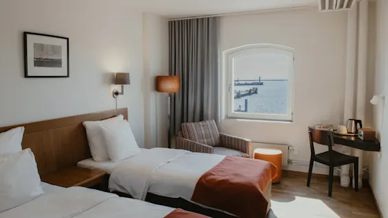 Viihtyisä hotellihuone, jossa kaksi sänkyä, työpöytä ja ikkuna kauniilla näkymällä merelle ja satamaan.