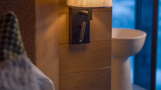 A wall-mounted lamp emits warm light over a wooden-paneled wall beside a neatly made bed in a cozy bedroom. A window with a blue-tinted outdoor view is visible.