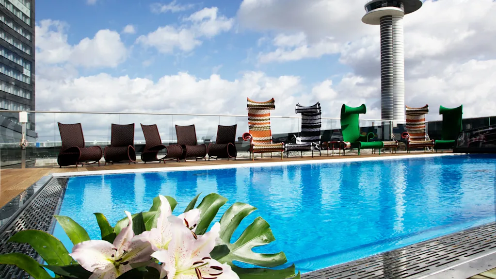 A rooftop swimming pool has blue water, surrounded by modern lounge chairs. A tall control tower is visible in the background under a partly cloudy sky.