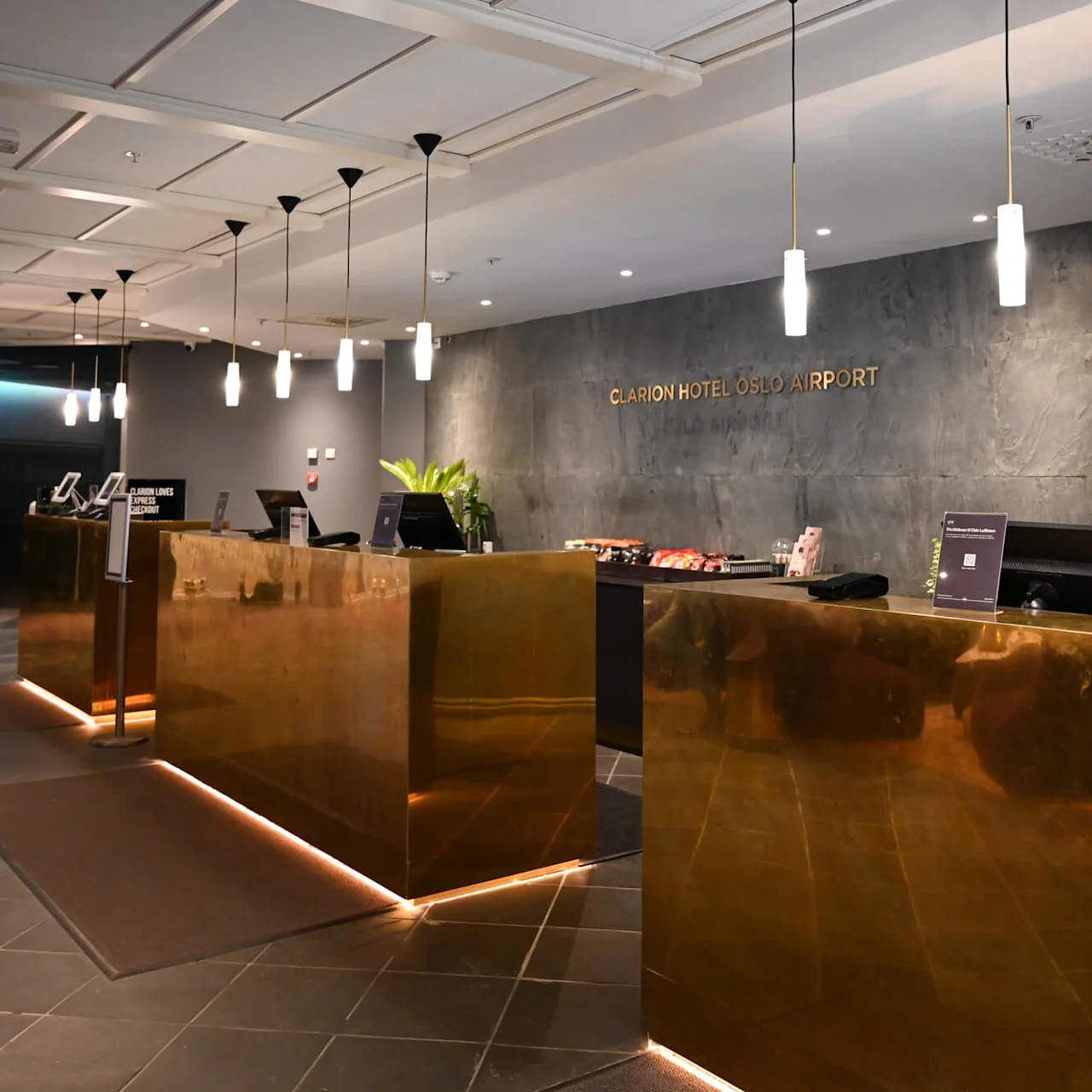 Reception med mässingsdiskar under pendellampor, en skylt med "Clarion Hotel Oslo Airport" på en texturerad vägg och modern inredning inklusive växter. Skyltar visar riktning till faciliteter.