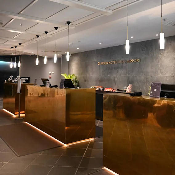 Reception area featuring brass counters beneath pendant lights, a sign reading "Clarion Hotel Oslo Airport" on a textured wall, and modern decor including plants. Signs indicate directions to facilities.