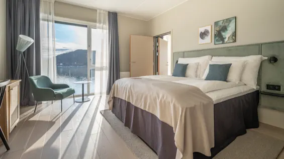 Lyst hotelværelse med stort vindue og naturskøn udsigt over sø og bjerge, med komfortabel seng og siddeområde.