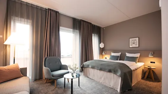 Kodikas hotellihuone, jossa on parivuode yöpöydän lampuilla, nojatuoli ja sohva. Huone on lämpimästi valaistu, ja verhot peittävät suuret ikkunat.