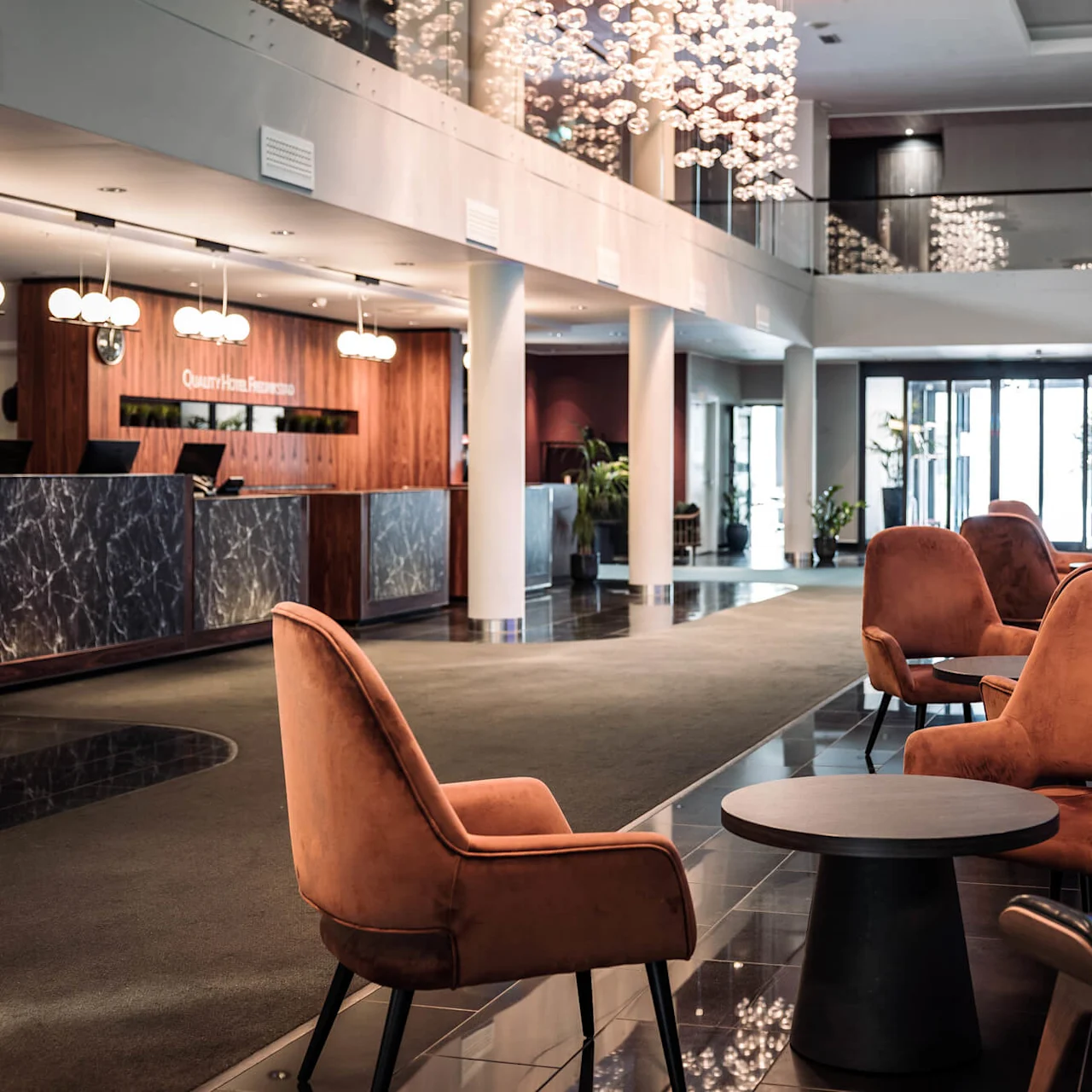 A reception desk with marble panels is attended and illuminated by modern lights; surrounded by plush chairs and tables in a spacious, stylish hotel lobby. Sign reads: "Quality Hotel Reception."