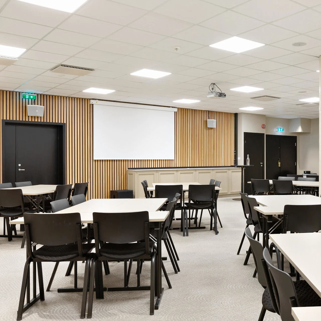 Konferencelokale med arrangerede borde og stole, inklusiv loftmonteret projektor og skærm. Beige vægge med træpaneler, mens sorte døre fører til tilstødende områder.