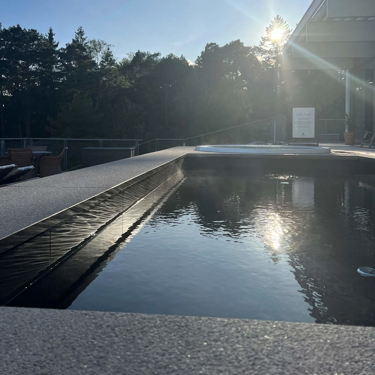 Pool reflecting sunlight, surrounded by a forest with tall trees. The sun is low in the sky, creating a serene atmosphere. A nearby sign reads “hidroterapija vrši…” (incomplete text).