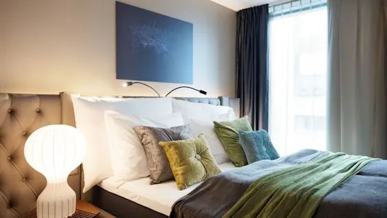 Bed adorned with cushions, covered by green and gray blankets, situated in a contemporary bedroom with soft lighting; artwork and lamp accentuate the calm ambiance.