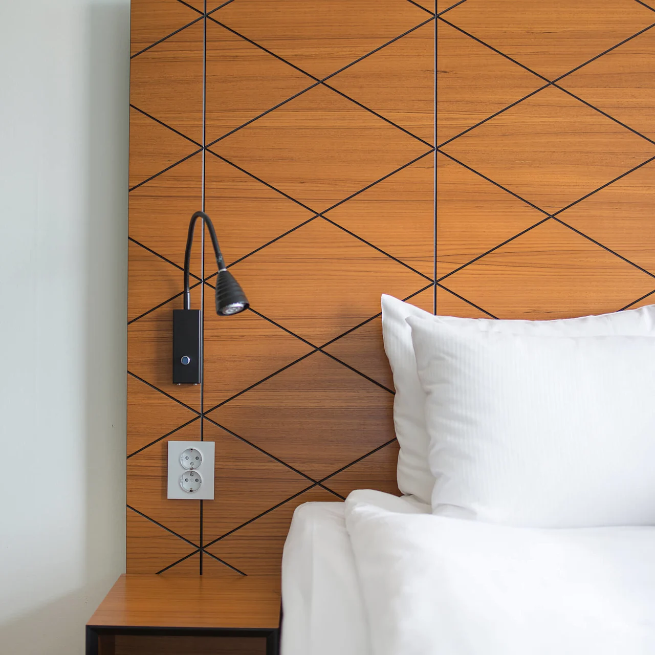A wooden headboard with geometric patterns features a black reading light and a power outlet above a bedside table. White bedding is partially visible.