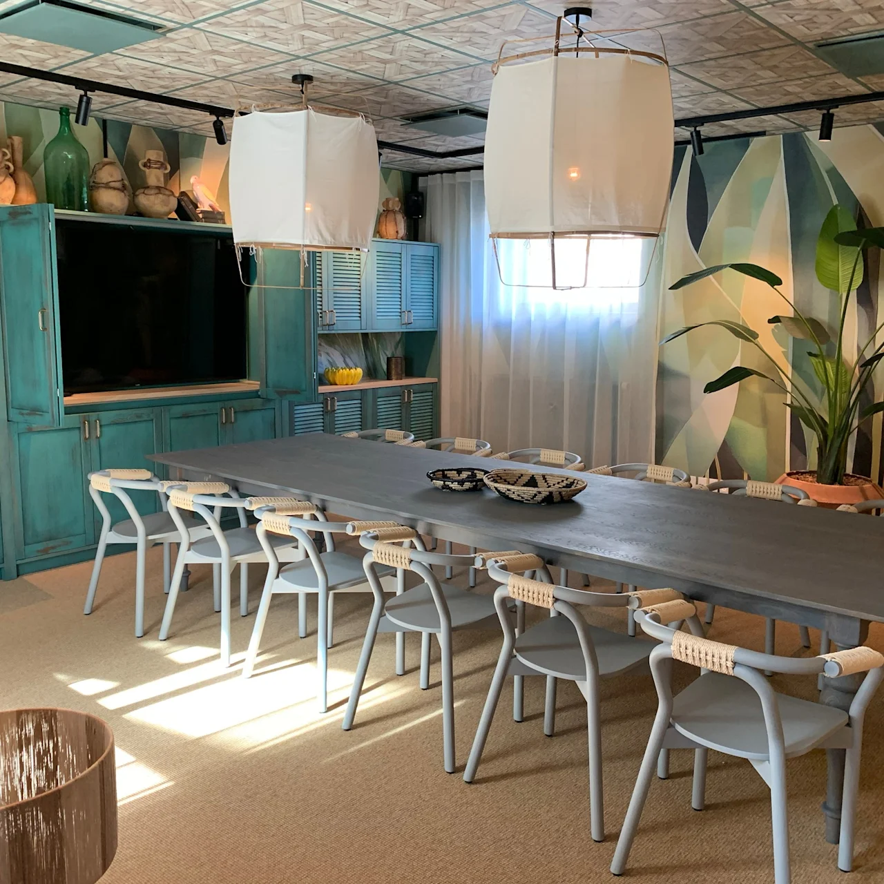 A long dining table is surrounded by white chairs, set under two large ceiling lamps. The room features turquoise cabinetry, abstract wall art, potted plants, and soft lighting.