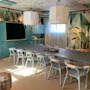 A long dining table is surrounded by white chairs, set under two large ceiling lamps. The room features turquoise cabinetry, abstract wall art, potted plants, and soft lighting.