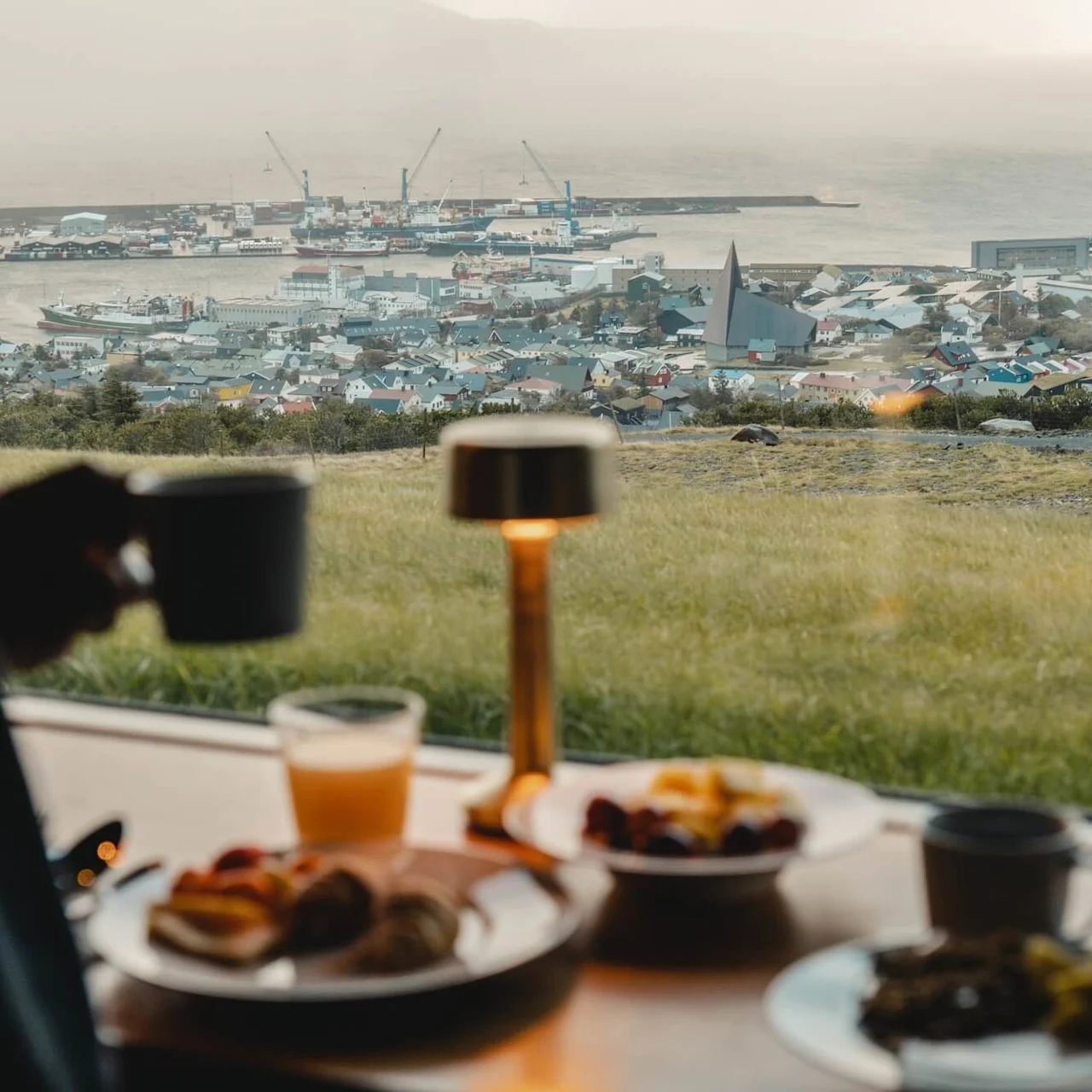 En person holder en kop over et morgenmadbord med tallerkener med mad og et glas juice. I baggrunden ses en kystby med farverige huse og fortøjede skibe.